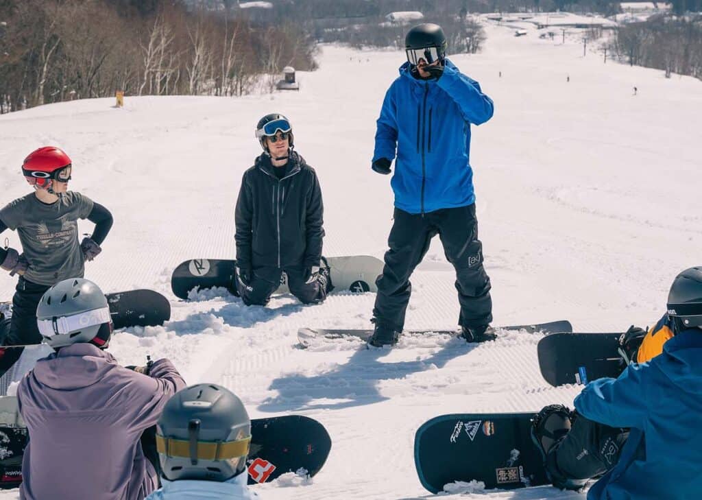 People getting a snowboard lesson. Sitting on the ski slope listening to the instructor.